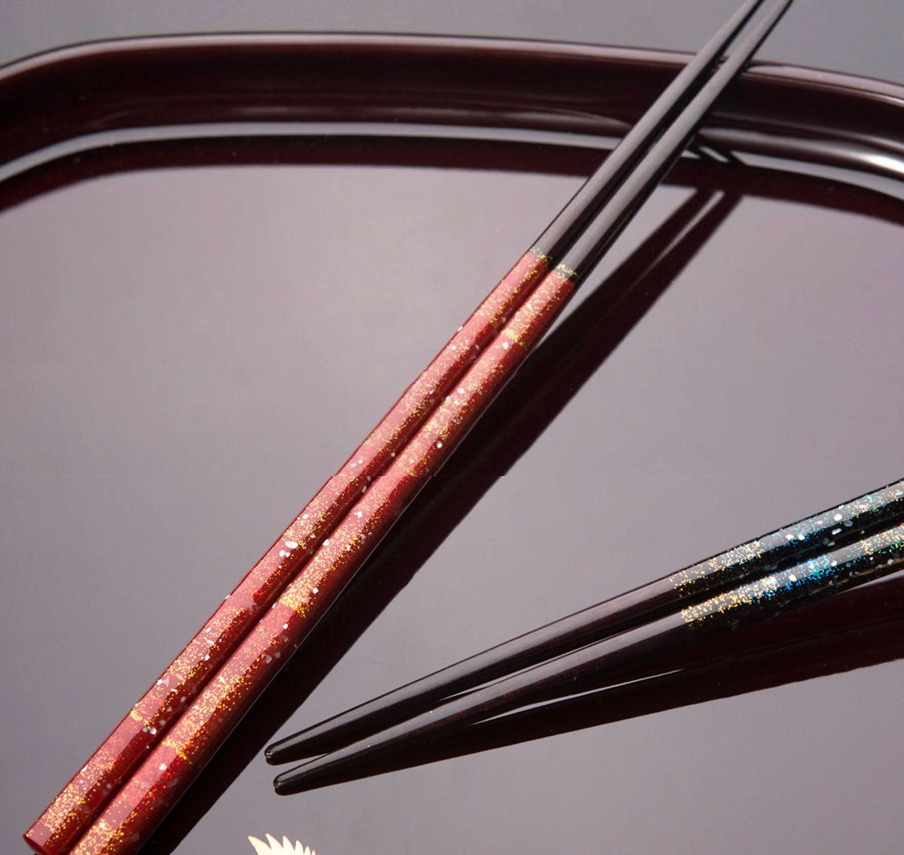 Red & blue Japanese handmade wooden chopsticks with gold & silver glitter on a dark tray.